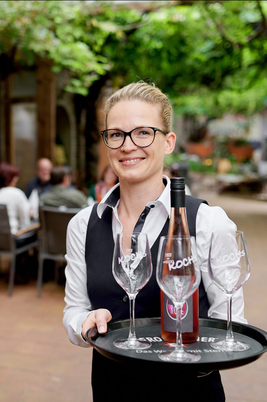 Freundliche Kellnerin mit Brille serviert Wein und Gläser auf einem Tablett in einem gemütlichen Außenbereich.